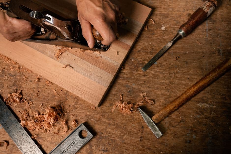 Hands using a hand plane on a striped glued-up board surrounded by chisels and shavings